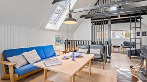 Ett rum i loftstil med högt i tak, synliga takbjälkar, ett takfönster, en blå soffa, ett träbord, en öppen spis och utsikt över omgivningarna.