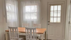 Un comedor con una mesa de madera y sillas blancas, una puerta blanca y una ventana con cortinas traslúcidas.
