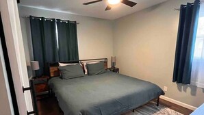 Un dormitorio con cama, ventilador de techo, mesita de noche, lámpara y ventana con cortinas.