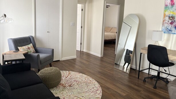 Una sala de estar moderna con un sillón gris, un taburete redondo con estampado, una mesa de centro de madera, una silla de oficina negra y un espejo grande en la pared.