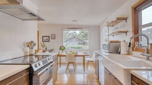 A modern kitchen with wooden cabinets, a central island, and a large window overlooking a house. - Four Bears by Avantstay Walk to Beach, Hot Tub, Ocean View (Arch Cape)