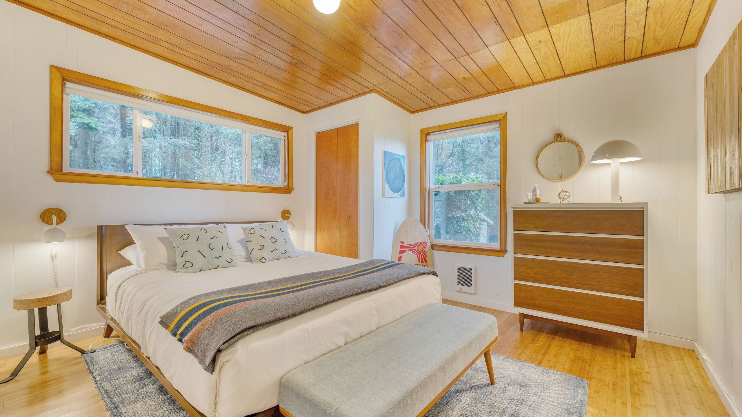 A bedroom with a wooden ceiling, large windows, a bed with patterned pillows, a bench, a dresser, and a nightstand with a lamp.