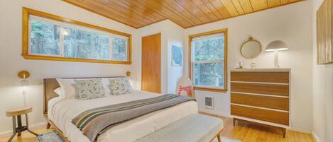 A bedroom with a wooden ceiling, large windows, a bed with patterned pillows, a bench, a dresser, and a nightstand with a lamp.