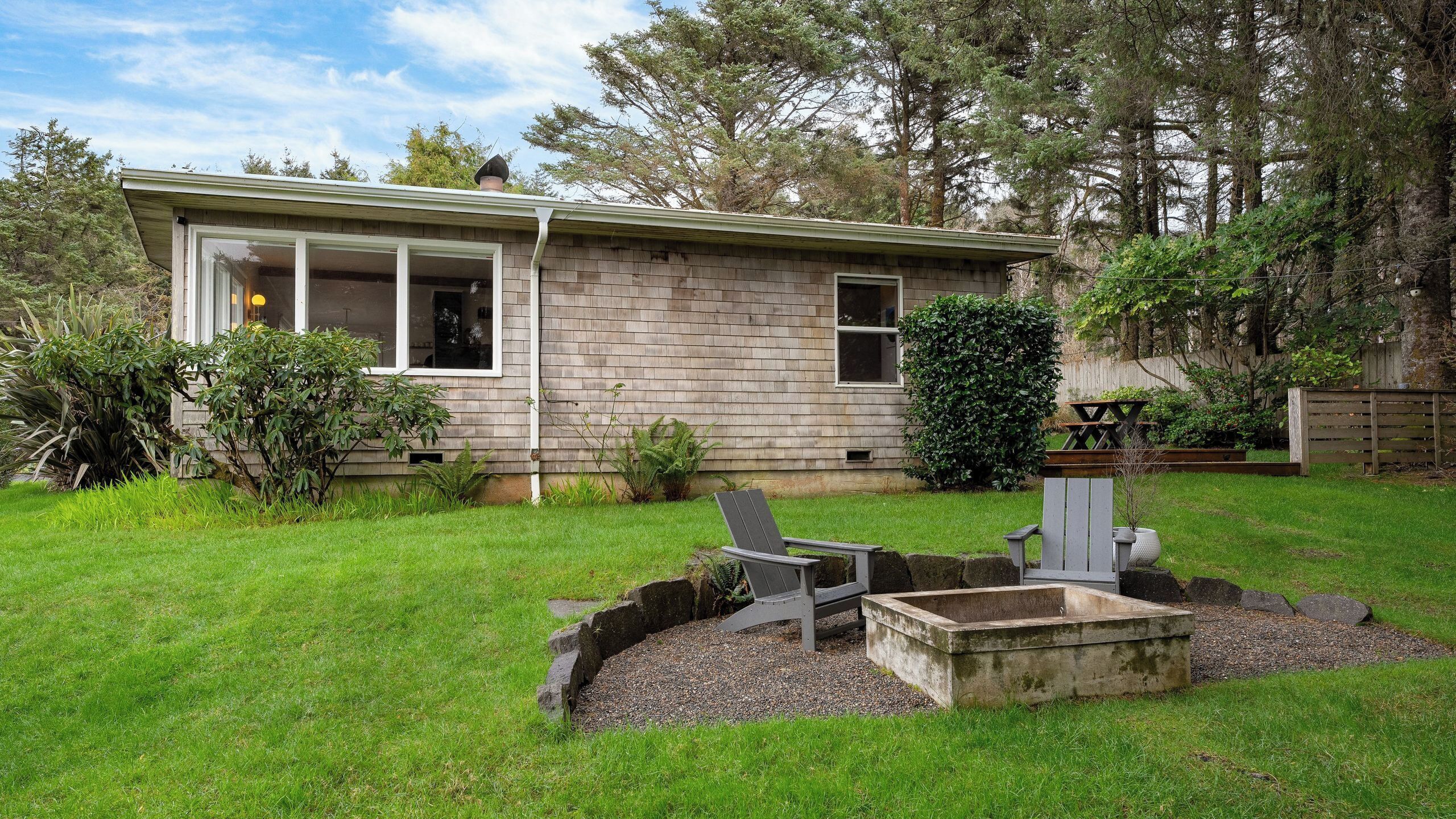A single-story house with a front porch, a small garden with a concrete planter and Adirondack chairs, and a wooden deck.