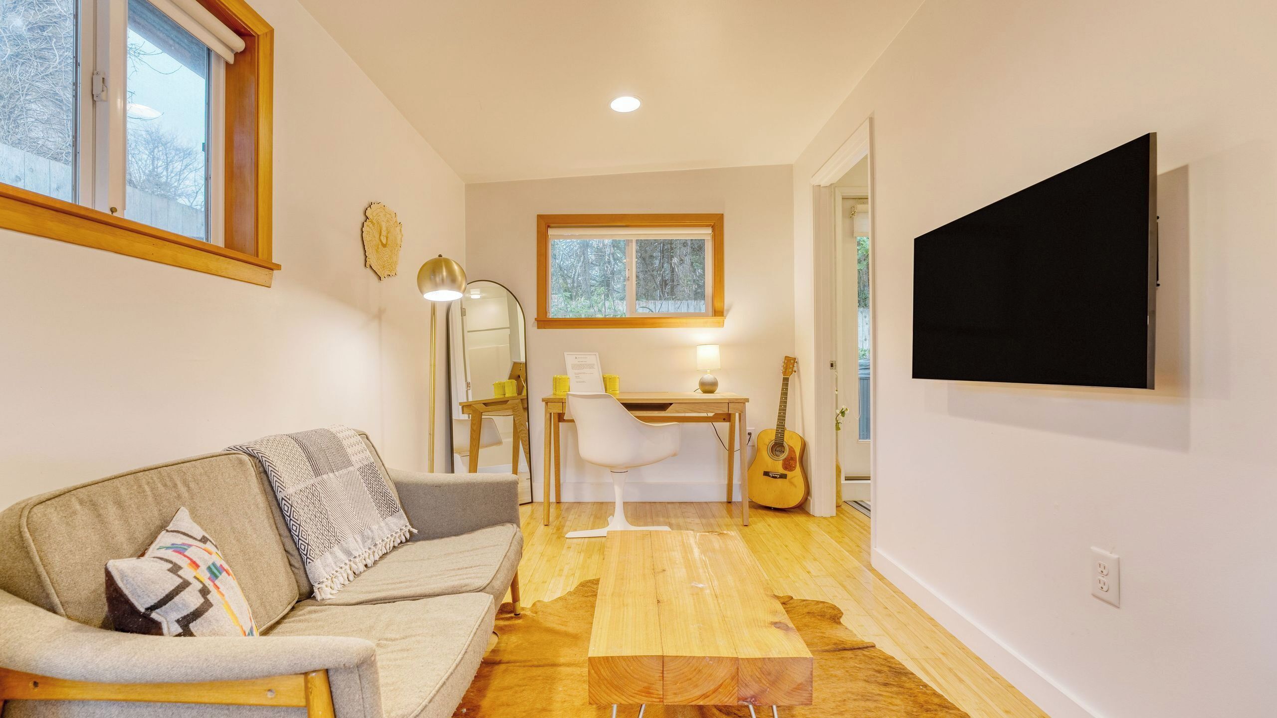 A cozy living room with a sofa, a desk, a guitar, and a TV mounted on the wall.