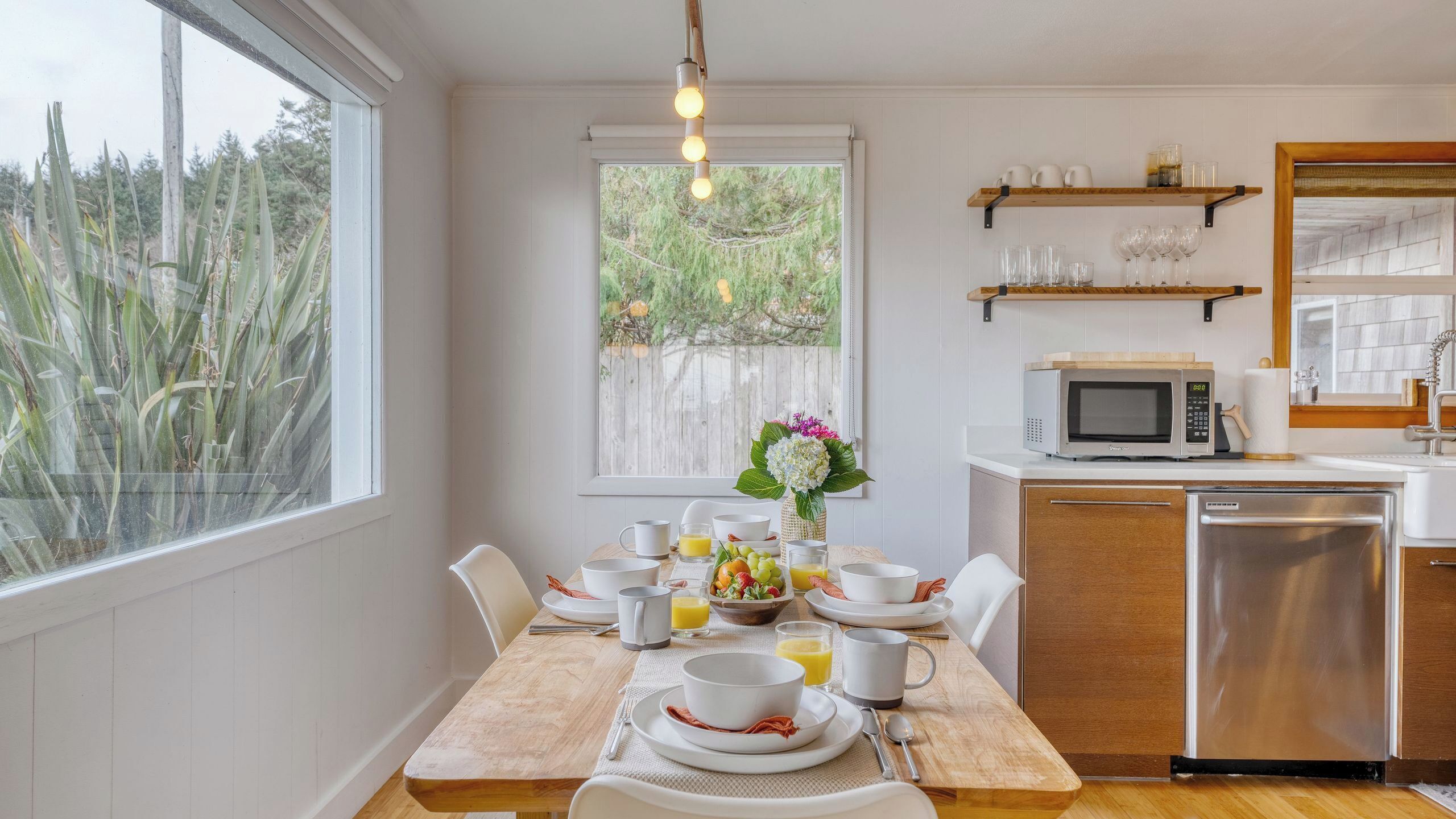 Una cocina moderna con comedor, que incluye una mesa de madera, sillas blancas, una encimera blanca, un microondas y un lavavajillas.