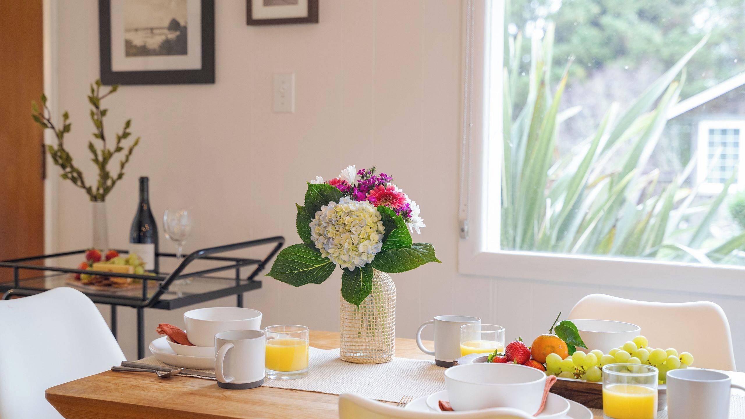 Una mesa de desayuno con un florero, fruta y alimentos para el desayuno.
