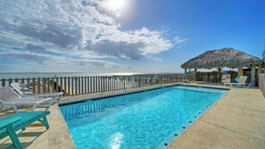 A pool with a wooden deck, lounge chairs, and a thatched-roof structure.