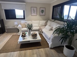 Ett vardagsrum med en vit soffa, ett soffbord med dekorativa föremål, en tv och ett stort fönster med persienner.