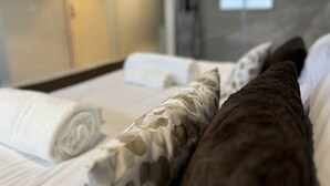 Habitación de hotel con cama, almohadas, una toalla enrollada, cabina de ducha de vidrio y una puerta de madera.