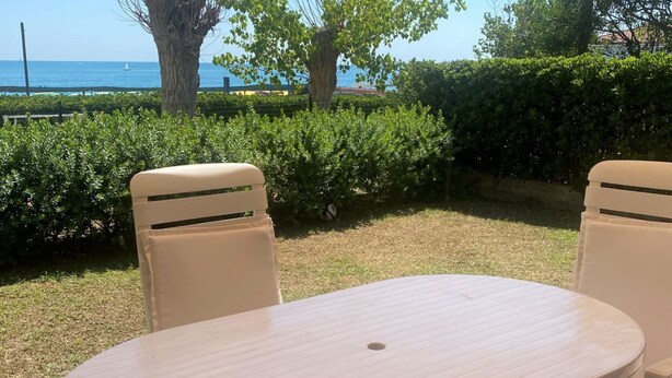 Ein Sitzbereich im Freien mit einem runden Tisch und zwei Stühlen, umgeben von Grünpflanzen und Blick auf das Meer.
