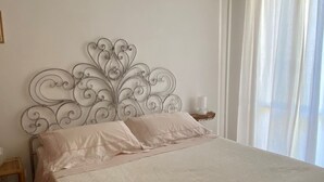 Ein ordentlich bezogenes Bett mit Metallkopfteil, ein Nachttisch mit Kerze und ein Fenster mit durchsichtigen Vorhängen.