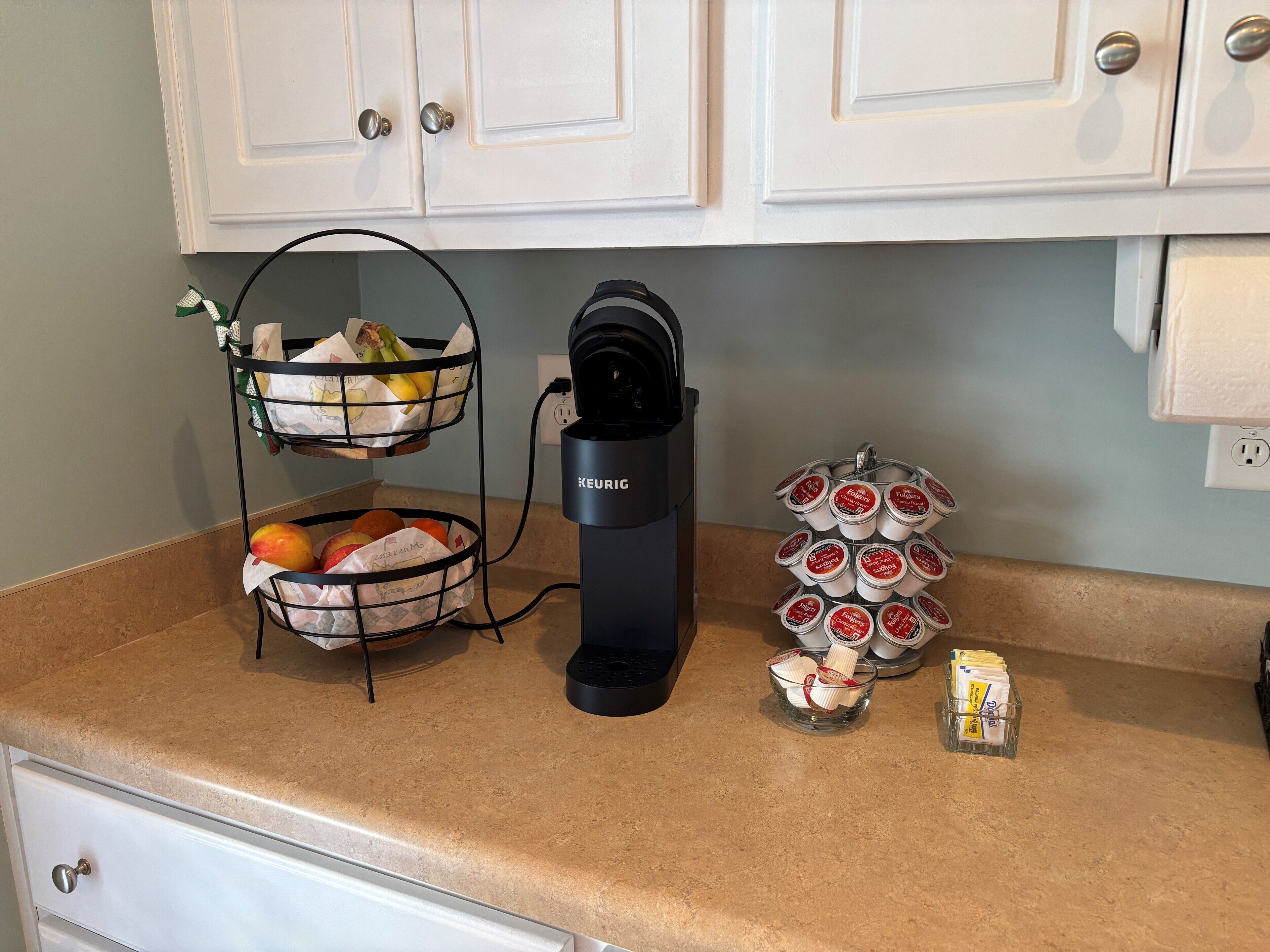 Una encimera de cocina con una cafetera Keurig, una cesta de fruta y un recipiente con cápsulas de café.