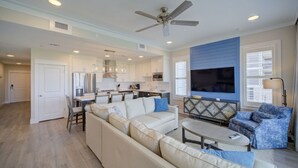 A modern living room with a large sofa, a coffee table, and a TV. There is a kitchen area with white cabinets and a dining table. - West-Facing Luxury Unit | Amazing Amenities (Destin)