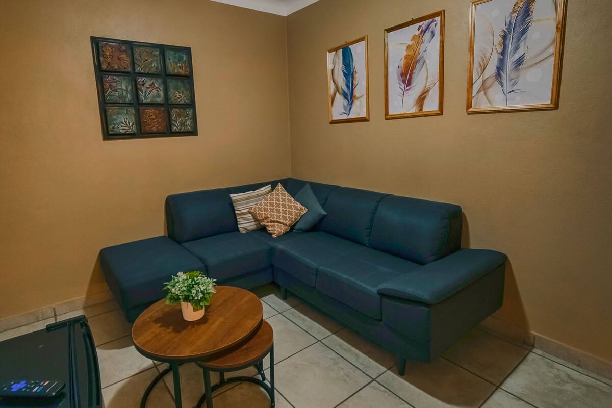 Een woonkamer met een hoekbank, een klein rond tafeltje met een plant, een plafondventilator en wanddecoratie in lijsten.