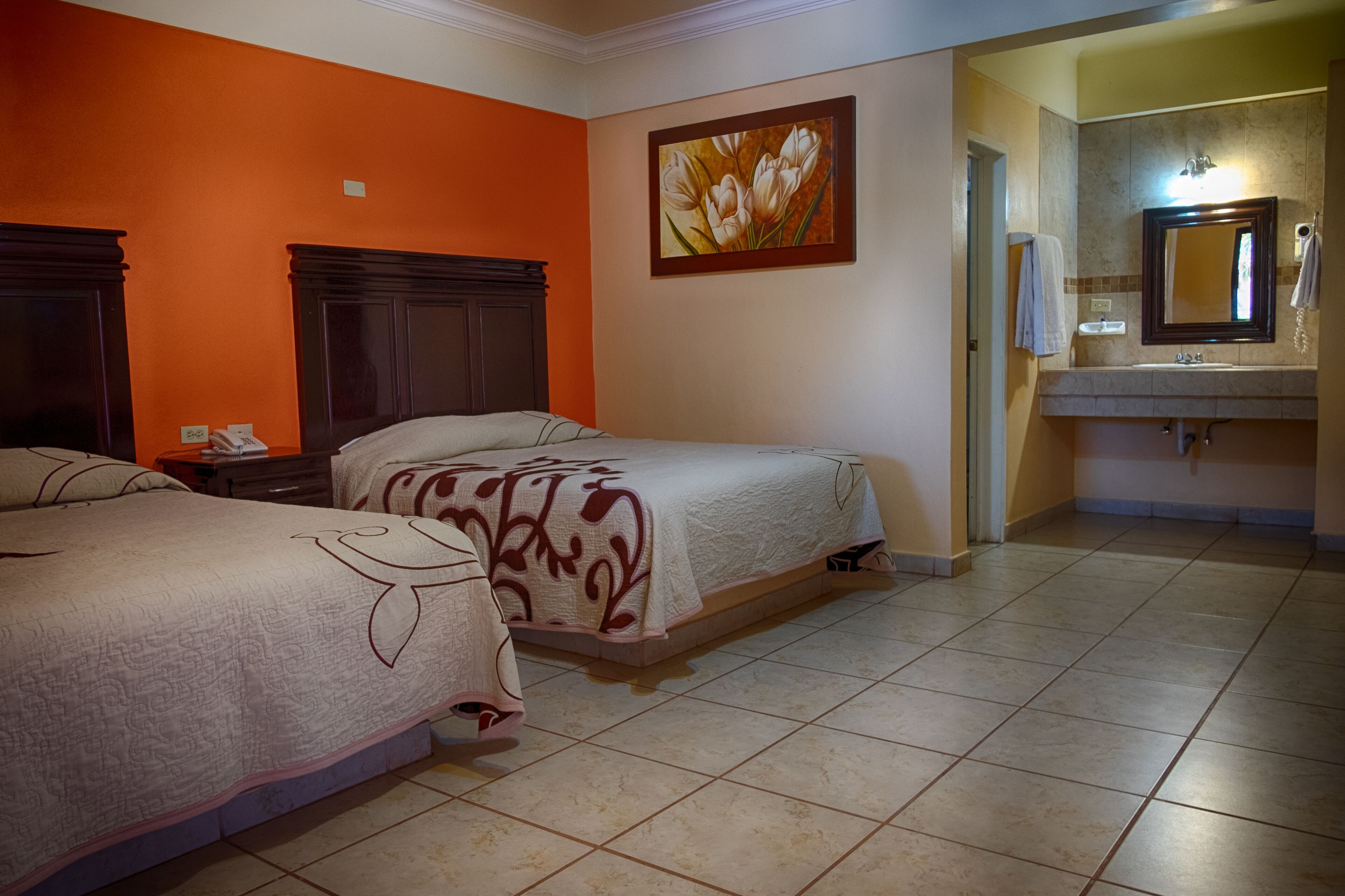 A hotel room with two beds, a bathroom with a sink and mirror, and a painting on the wall.