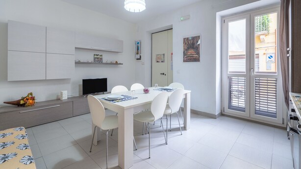 Een moderne keuken met witte kastjes, een eettafel met stoelen, een televisie en een raam met jaloezieën.
