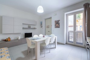 A modern kitchen with white cabinets, a dining table with chairs, a television, and a window with blinds.