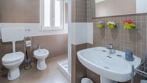 Een badkamer met toilet, bidet en wastafel, met een raam en een plankje met bloemen.