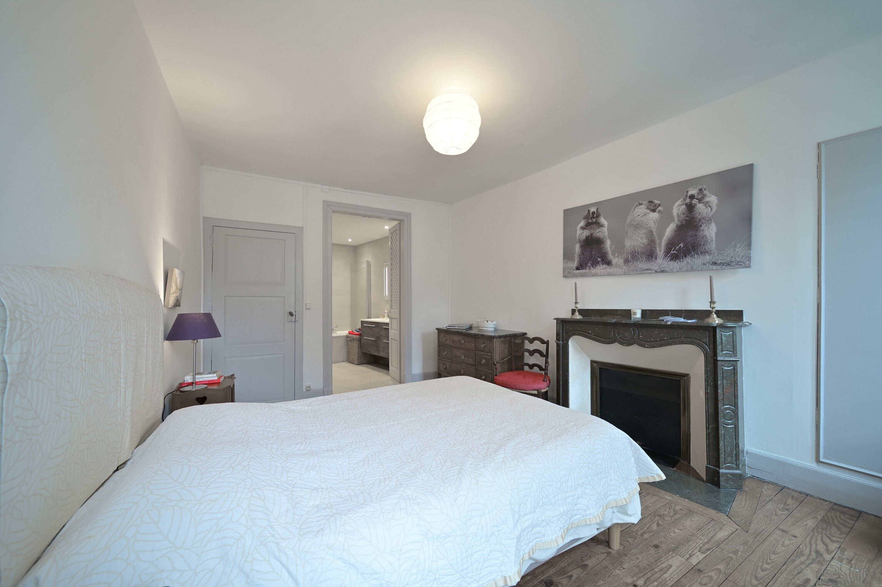 A bedroom with a large bed, a fireplace, a door leading to another room, and a wall-mounted painting.
