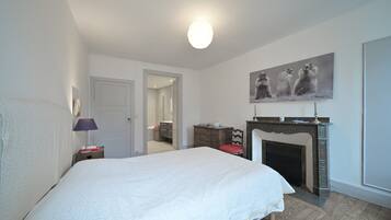A bedroom with a large bed, a fireplace, a door leading to another room, and a wall-mounted painting.