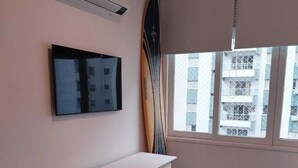 A room with a white cabinet, a mounted TV, a surfboard leaning against the wall, a window with a view of buildings, and a black rug. - Você Ficará Hospedado a Menos de 5min a pé da Praia de Pitangueiras (Centro Guarujá)