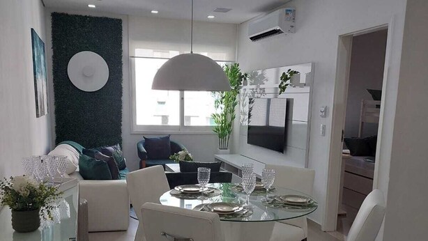 A modern living room with a glass dining table, white sofas, and a large TV. - Você Ficará Hospedado a Menos de 5min a pé da Praia de Pitangueiras (Centro Guarujá)