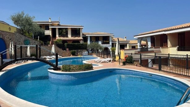 A swimming pool with lounge chairs, a stone wall, and buildings with tiled roofs. - Apartment 'Dimora Frau' with Shared Pool, Wi-Fi and Air Conditioning (Villasimius)