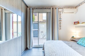 A bedroom with a bed, a sliding glass door to a balcony, a small table with a lamp, and a wall-mounted shelf with decorative items. - Holiday Home “Villa Almina” Near Naturist Area (Agde)