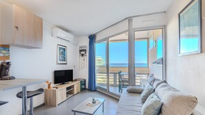A modern living room with a flat-screen TV, a wooden cabinet, a coffee machine, a dining table with bar stools, a sofa with cushions, and a large sliding glass door leading to a balcony. - Studio Apartment 'Vue Mer' with Sea View, Wi-Fi and Air Conditioning (Canet-en-Roussillon)