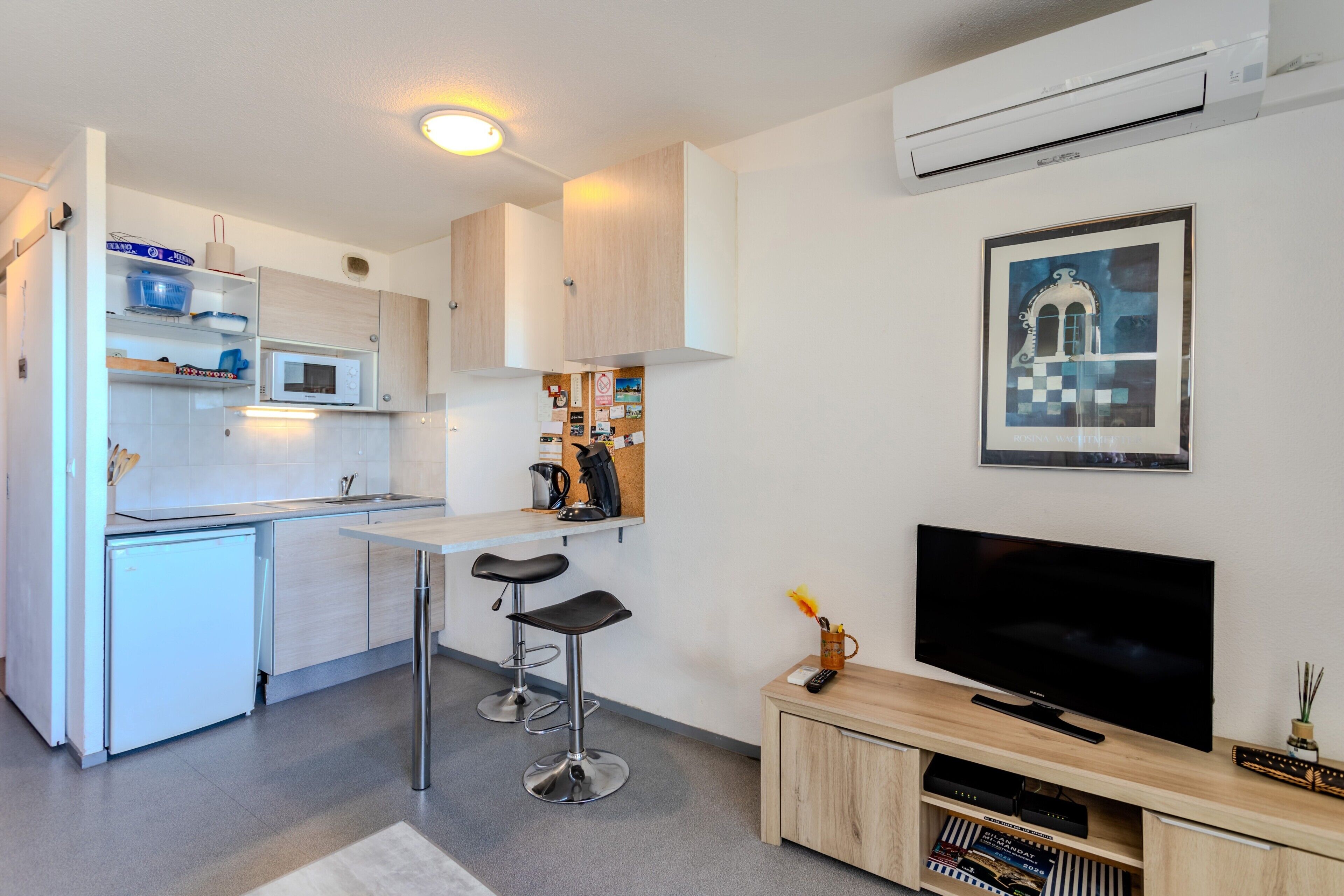 Cocina compacta con microondas, lavamanos y taburetes de bar. Se ve un televisor de pantalla plana y un mueble pequeño.