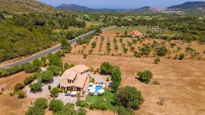A large house with a swimming pool, surrounded by a dry field and trees, with a road and hills in the background.