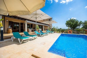 Un espace aménagé au bord de la piscine, avec des chaises longues et des parasols, une piscine à l’eau d’un bleu limpide et un bâtiment doté d’un toit recouvert de tuiles.