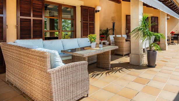 A covered outdoor seating area with wicker furniture, a table, and potted plants.
