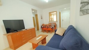 A living room with a blue sofa, a wooden TV stand, a dining table with chairs, and a mirror above the table. - ACV - Playa Coral I-2ª Linea Planta 3 Norte (OROPESA)