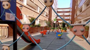 A playground with swings and slides, surrounded by a brick building and potted plants. - ACV - Playa Coral I-2ª Linea Planta 3 Norte (OROPESA)