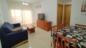 A living room with a sofa, a TV on a wooden cabinet, a dining table with chairs, and a door. - ACV - Playa Coral I-2ª Linea Planta 3 Norte (OROPESA)