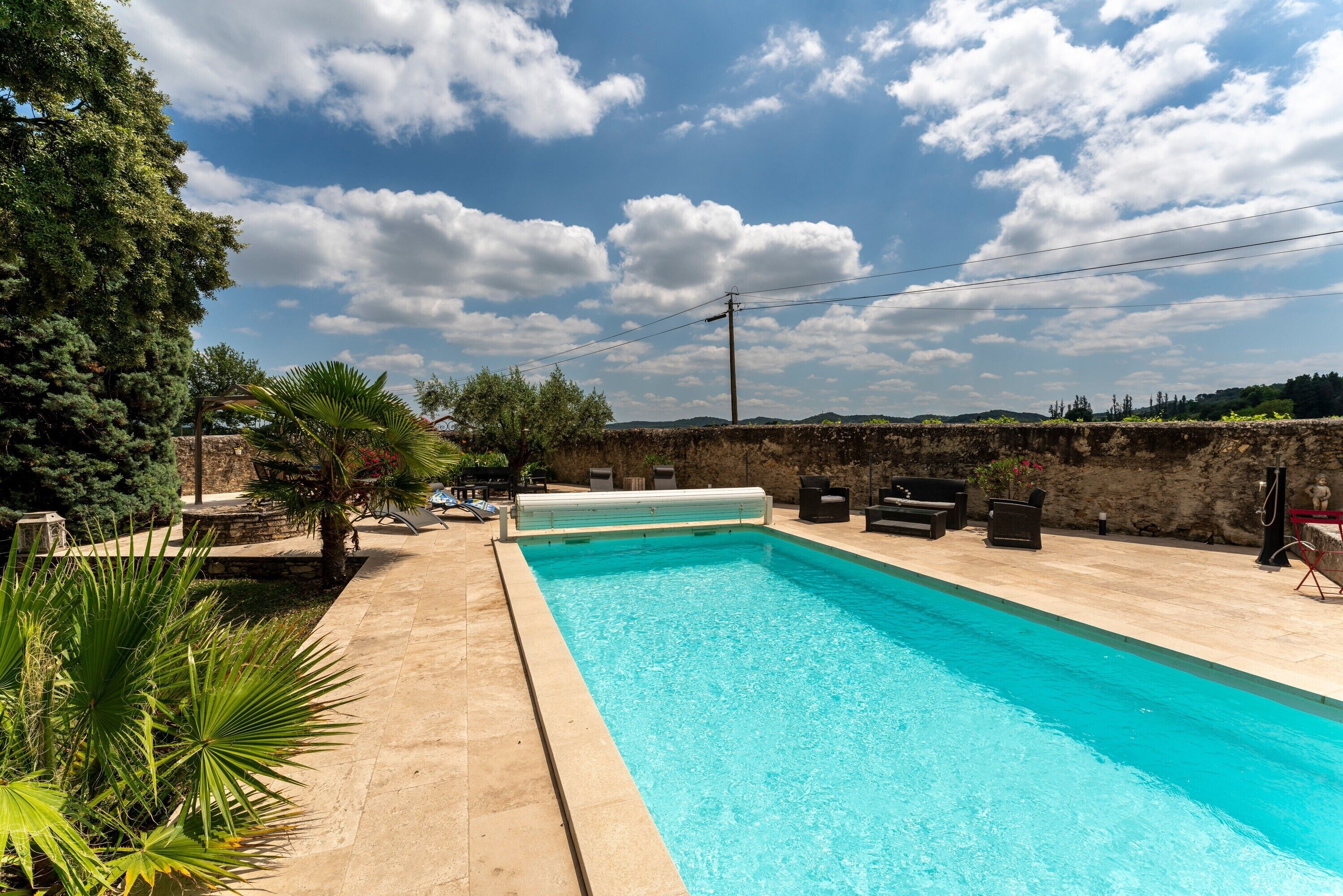 Una piscina rectangular amb una zona enrajolada, envoltada de vegetació i un mur de pedra.