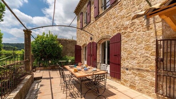 Un edificio de piedra con un patio empedrado, una mesa lista para comer y contraventanas rojas.