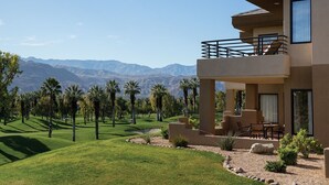 Un edificio moderno de dos pisos con balcón, un patio con mobiliario exterior y vistas a un campo de golf y montañas.