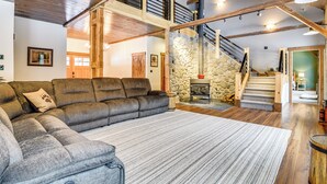A spacious living area with a large sectional sofa, a stone fireplace, and a wooden staircase leading to an upper level. - Maine Home w/ Hot Tub & Snowmobile Trail Access! (Township of Milton)