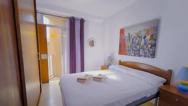Una habitación de hotel con una cama, una mesita de noche, un cuadro en la pared y una puerta que da a otra habitación.