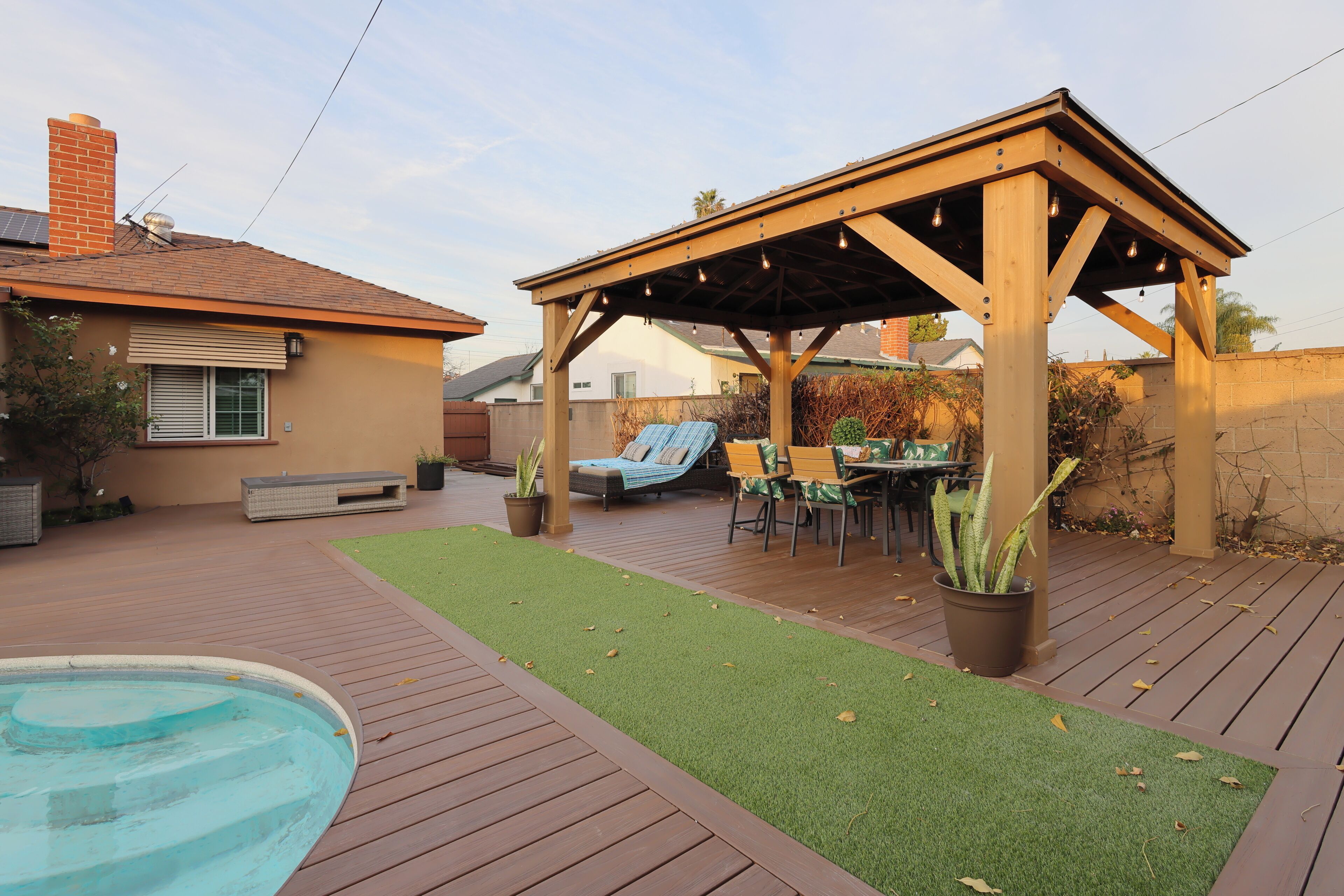 A backyard with a wooden deck, a pool, a covered patio area, and outdoor furniture.