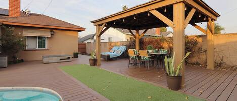 A backyard with a wooden deck, a pool, a covered patio area, and outdoor furniture.
