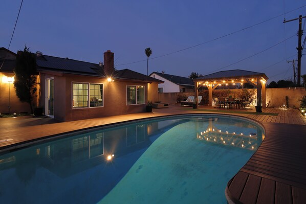 A swimming pool with a wooden deck, a covered patio with string lights, and a house with solar panels. - Perfect Home Near Disneyland (Anaheim)