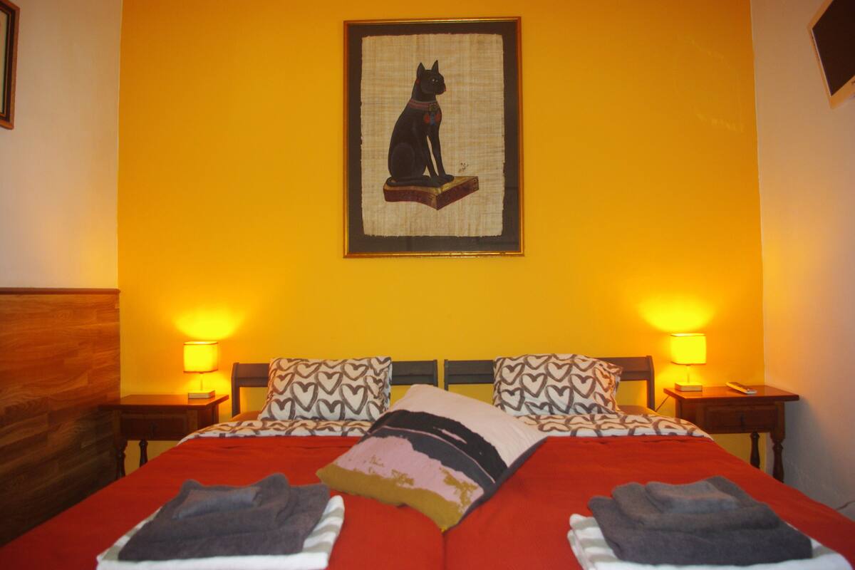 A neatly made bed with a red bedspread, two heart-patterned pillows, and a framed artwork of a cat on the wall.