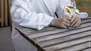Una persona con una veste bianca seduta a un tavolo di legno all'aperto, con un bicchiere di vino bianco, con vista sulla città.