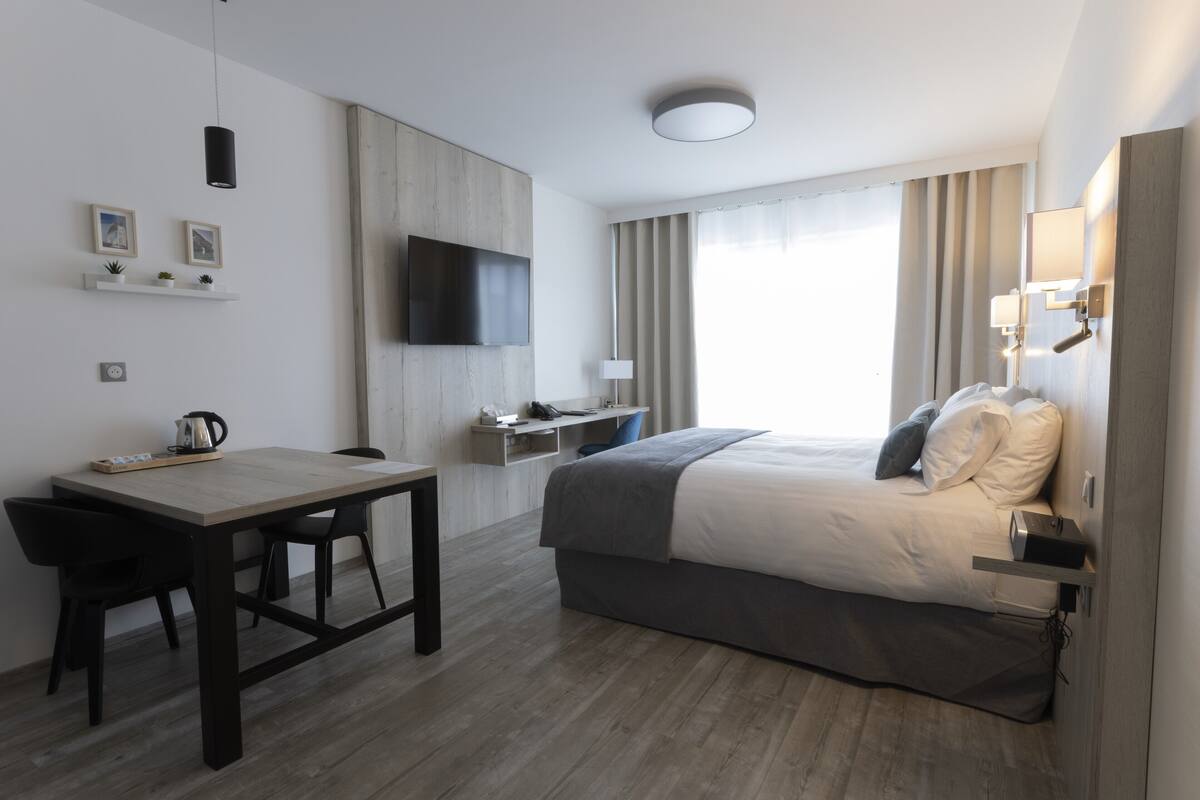 A modern hotel room with a bed, desk, chairs, a TV, and a window with curtains.