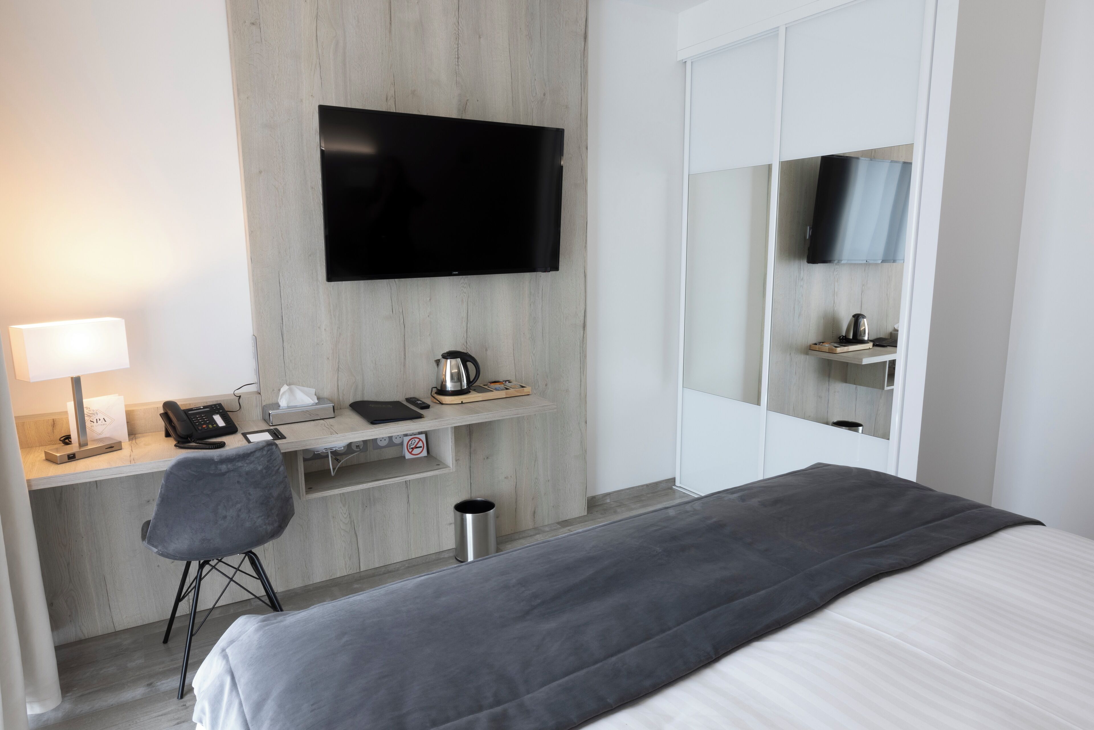 A modern hotel room with a bed, a desk with a chair, a flat-screen TV mounted on the wall, a mirror, and a small shelf with a kettle.