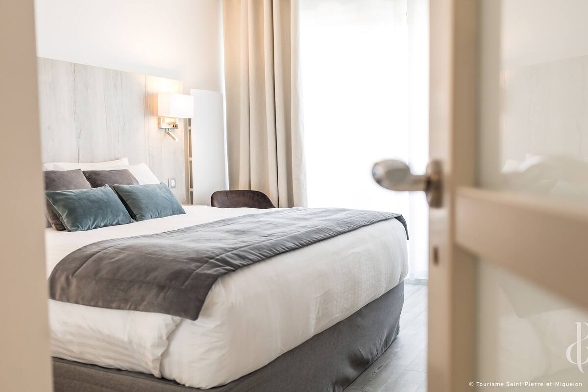A hotel room with a bed, a nightstand, a lamp, and a view of the outdoors.
